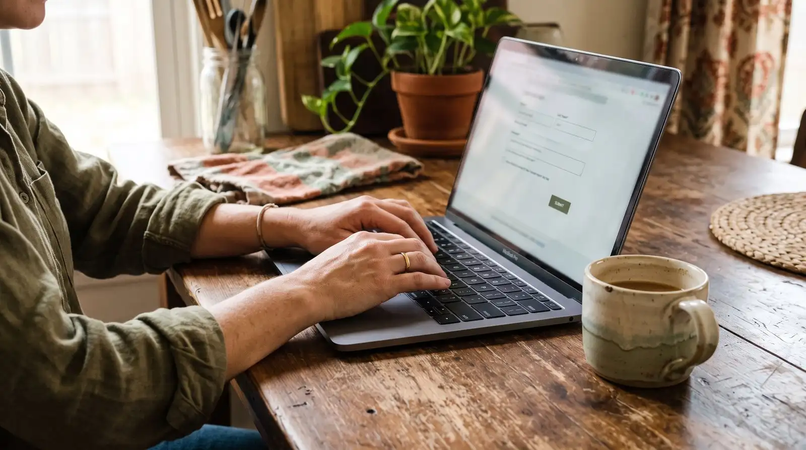 Hands typing on a laptop at a wooden kitchen table beside a coffee mug